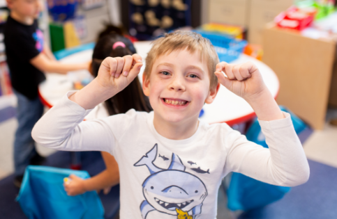 A young child in a classroom setting smiling and rasining his hands up.